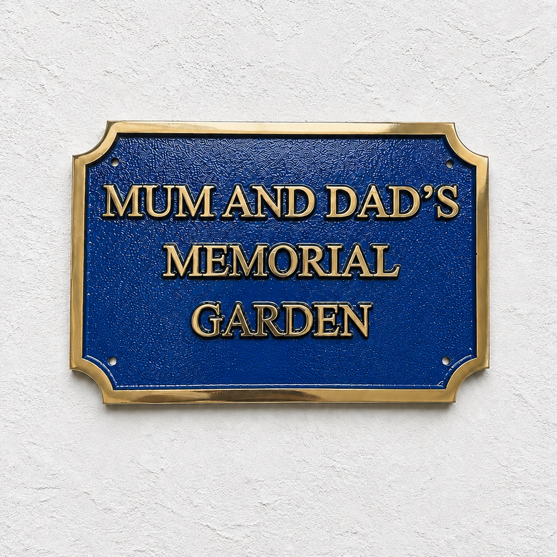 A rectangular blue memorial garden sign with gold lettering reading 'MUM AND DAD'S MEMORIAL GARDEN' and a gold border, mounted on a textured white wall.
