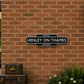 A decorative black and white sign reading 'HENLEY ON THAMES RAILWAYS' mounted on a red brick wall, with a vintage train station platform, stacked brown trunks on a cart, and a planter with flowers nearby.