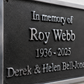 A close-up of a memorial plaque with raised silver lettering on a dark metal background, mounted on a light textured wall. The plaque reads: 'In memory of Roy Webb 1936 - 2025' and 'Derek & Helen Bell-Jones'.