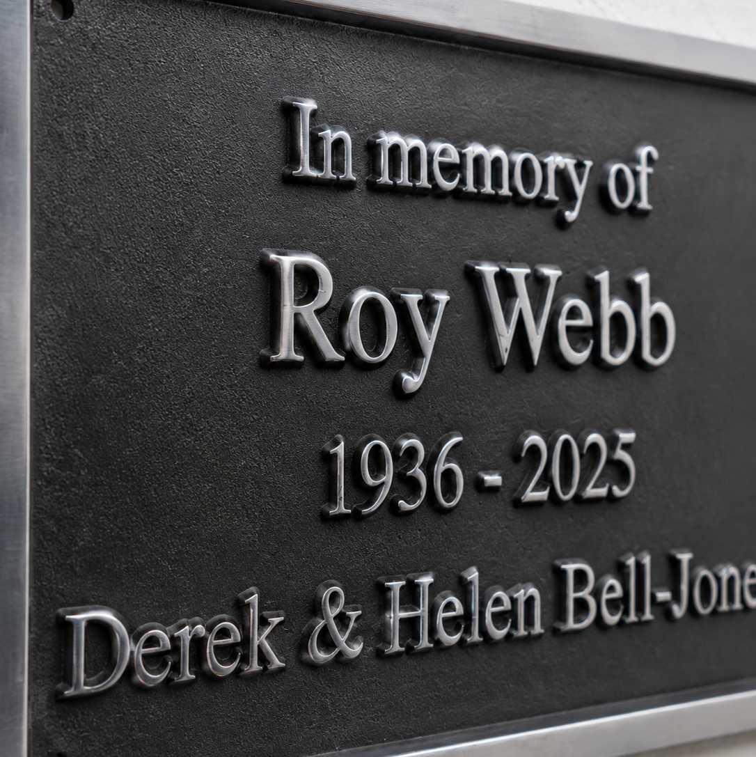 A close-up of a memorial plaque with raised silver lettering on a dark metal background, mounted on a light textured wall. The plaque reads: 'In memory of Roy Webb 1936 - 2025' and 'Derek & Helen Bell-Jones'.