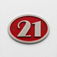 An oval house number plaque featuring the number 21 in raised silver lettering on a textured red background, mounted on a white textured wall.