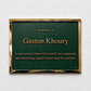 A rectangular green memorial plaque with a polished gold-toned metal border, mounted on a textured white wall. The plaque features raised gold lettering that reads 'In memory of Gaston Khoury' and a commemorative quote below.