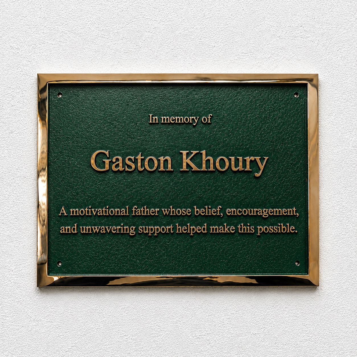 A rectangular green memorial plaque with a polished gold-toned metal border, mounted on a textured white wall. The plaque features raised gold lettering that reads 'In memory of Gaston Khoury' and a commemorative quote below.