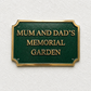 A rectangular green memorial garden plaque with gold lettering reading 'MUM AND DAD'S MEMORIAL GARDEN' and a gold border, mounted on a textured off-white wall.
