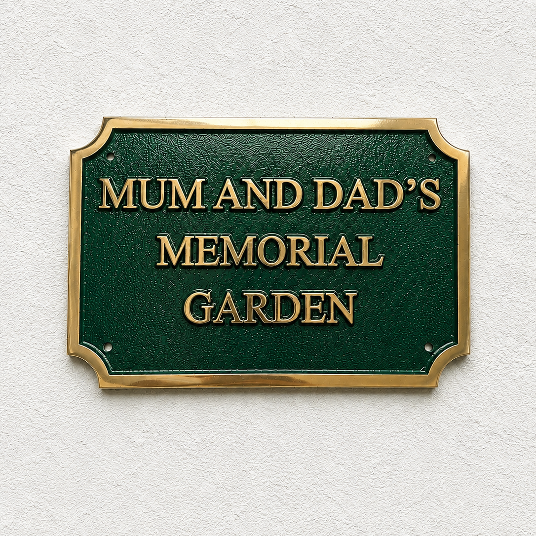 A rectangular green memorial garden plaque with gold lettering reading 'MUM AND DAD'S MEMORIAL GARDEN' and a gold border, mounted on a textured off-white wall.