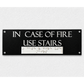 A rectangular black emergency exit sign with white text reading 'IN CASE OF FIRE USE STAIRS' and a white Braille section below, mounted on a textured white wall.