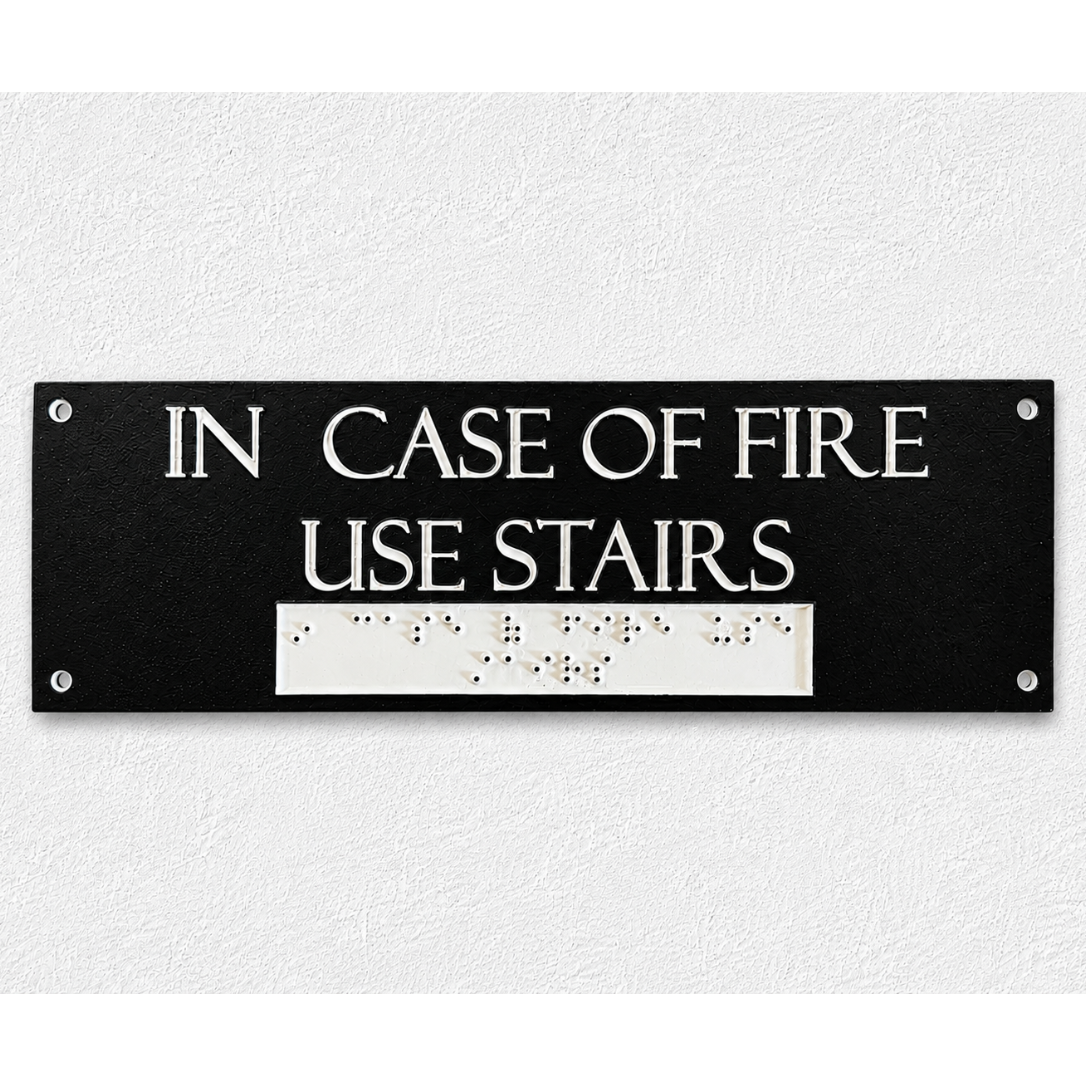 A rectangular black emergency exit sign with white text reading 'IN CASE OF FIRE USE STAIRS' and a white Braille section below, mounted on a textured white wall.