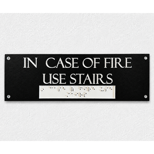 A rectangular black emergency exit sign with white text reading 'IN CASE OF FIRE USE STAIRS' and a white Braille section below, mounted on a textured white wall.