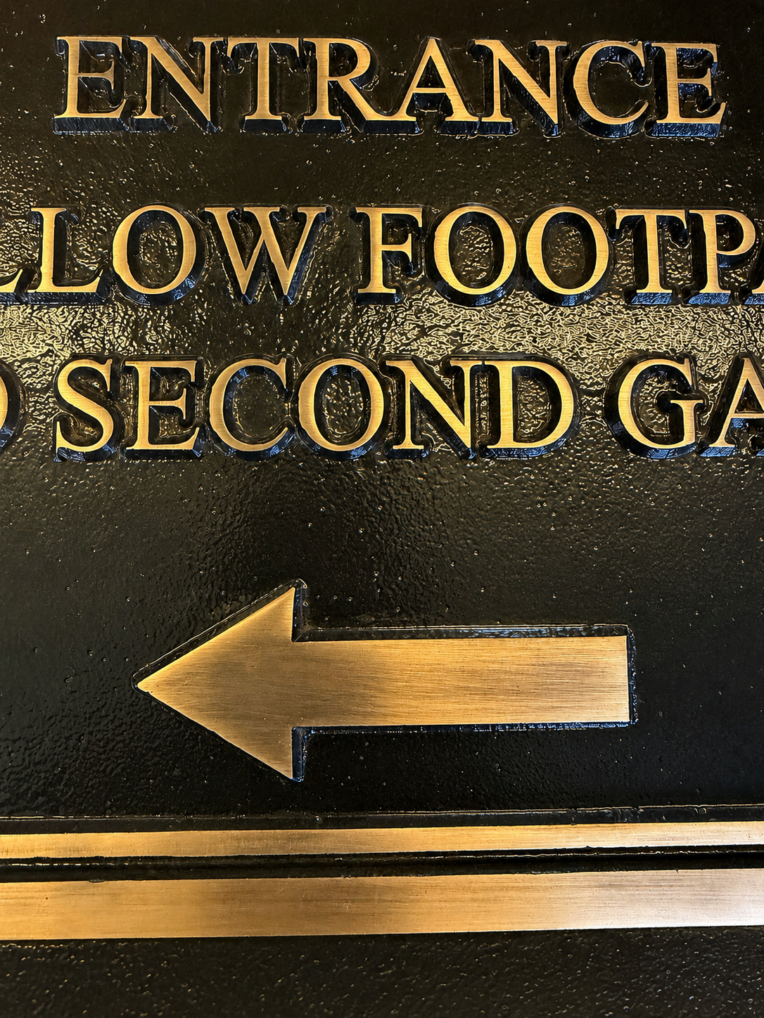 Close-up of a black metal sign with raised gold lettering reading 'ENTRANCE' and 'SECOND GA.