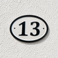 An oval house number plaque with the number 13 in black on a white background, mounted on a textured white stucco wall.