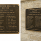 Two bronze commemorative headstone plaques for Pembroke College Oxford displayed side by side against a light beige stone wall. The left plaque is fully visible and shows the front with engraved names and coaches.