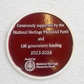 A round dark red commemorative plaque mounted on a textured white wall. The plaque features white text acknowledging support from the National Heritage Memorial Fund and UK government funding for the years 2023-2026.