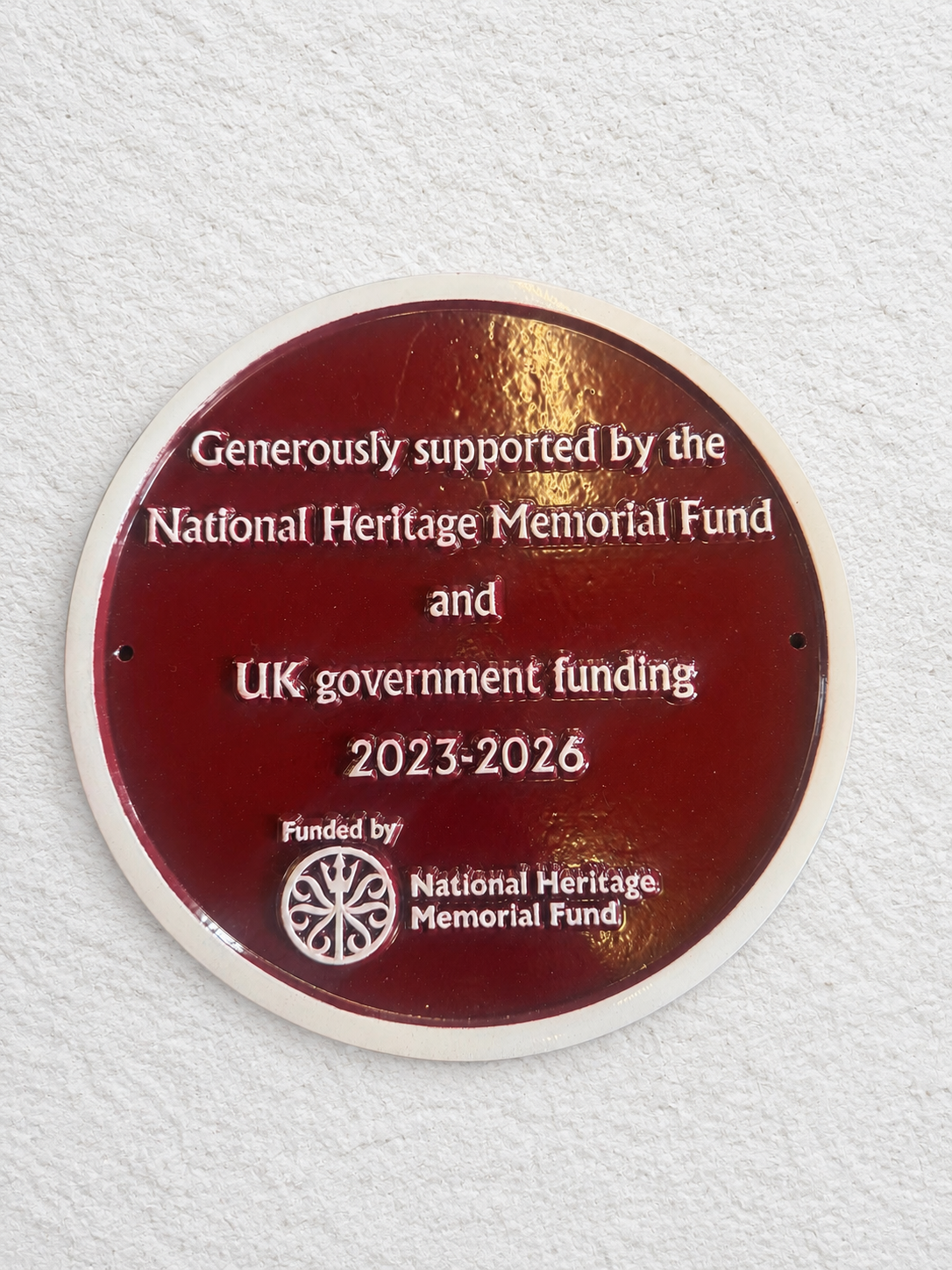 A round dark red commemorative plaque mounted on a textured white wall. The plaque features white text acknowledging support from the National Heritage Memorial Fund and UK government funding for the years 2023-2026.