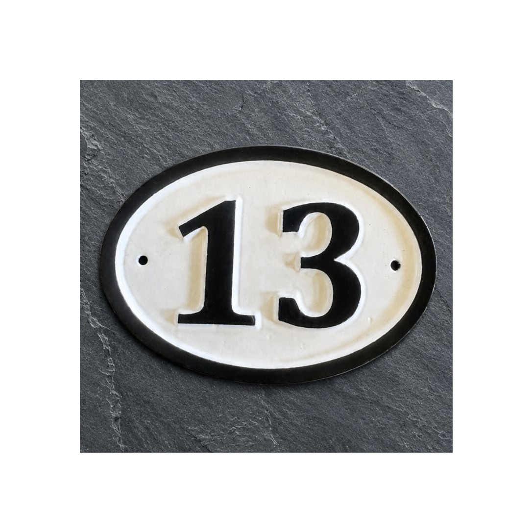 An oval house number plaque displaying '13' in black on a cream background with a black border, mounted on a dark gray slate-textured wall.