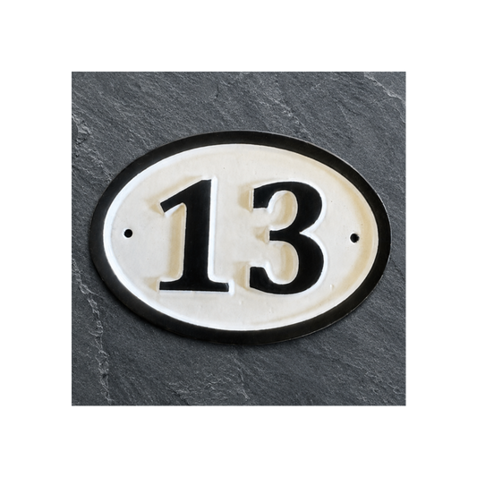 An oval house number plaque displaying '13' in black on a cream background with a black border, mounted on a dark gray slate-textured wall.