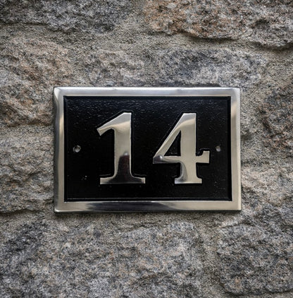 House number '14' on a black plaque with silver border mounted on a stone wall.