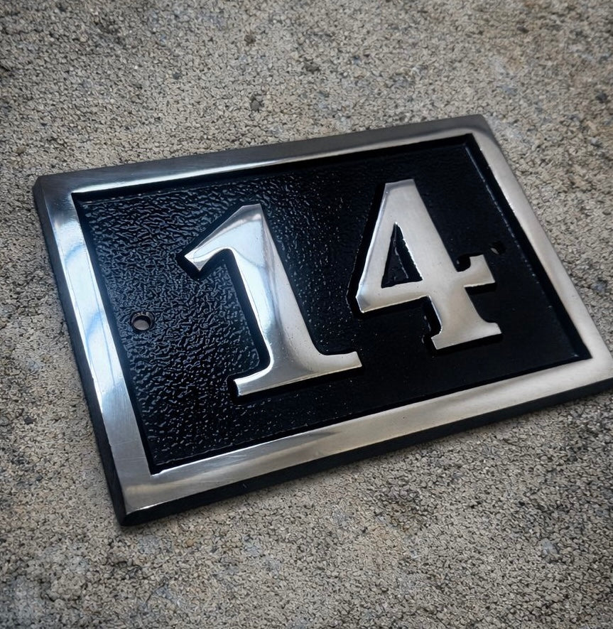 House number '14' on a black plaque with silver border, placed on concrete.
