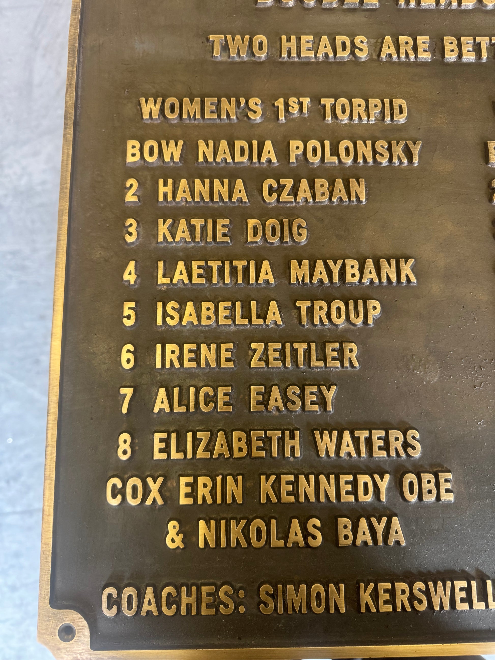 A close-up of a bronze or dark metal commemorative plaque with raised gold-colored text listing women's tennis players and coaches.