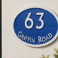 An oval blue and white house number sign reading '63 GRIFFIN ROAD' mounted on a textured white stucco wall, with green foliage partially framing the scene.