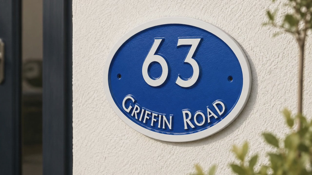 An oval blue and white house number sign reading '63 GRIFFIN ROAD' mounted on a textured white stucco wall, with green foliage partially framing the scene.