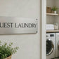 A rectangular brushed metal sign with the words 'GUEST LAUNDRY' in black serif lettering, mounted on a white textured wall.
