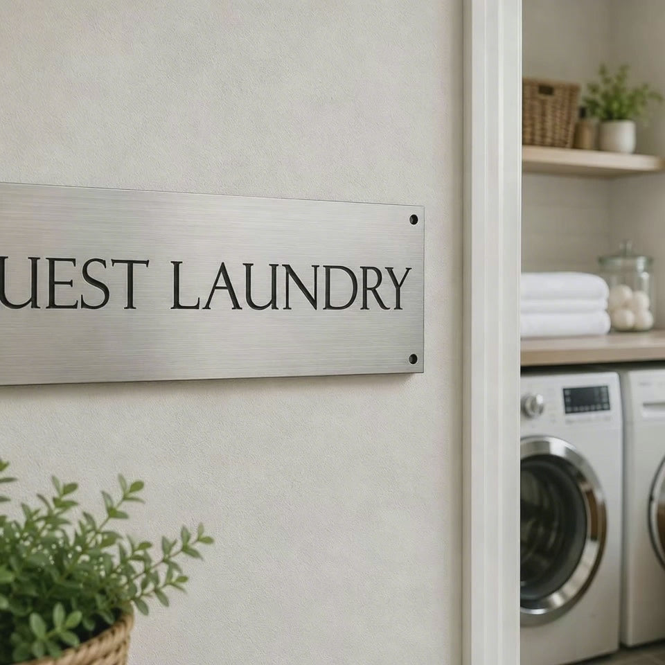 A rectangular brushed metal sign with the words 'GUEST LAUNDRY' in black serif lettering, mounted on a white textured wall.