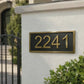 A rectangular black house number plaque with gold numerals and a gold border, mounted on a white stucco wall next to a black gate and green shrubs.
