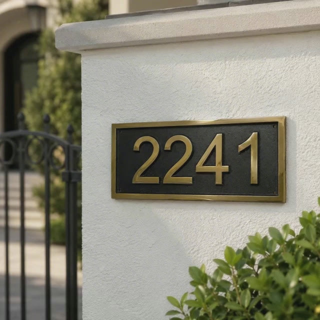A rectangular black house number plaque with gold numerals and a gold border, mounted on a white stucco wall next to a black gate and green shrubs.