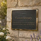 A rectangular black memorial plaque with a polished gold-toned border, mounted on a textured white wall. The plaque reads 'In memory of Gaston Khoury' with additional commemorative text below.