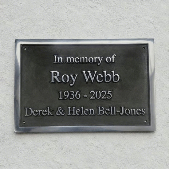 A rectangular dark metal memorial plaque mounted on a textured white wall, with raised silver text reading 'In memory of Roy Webb 1936 - 2025 Derek & Helen Bell-Jones'.
