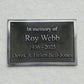 A rectangular dark metal memorial plaque mounted on a textured white wall, with raised silver text reading 'In memory of Roy Webb 1936 - 2025 Derek & Helen Bell-Jones'.