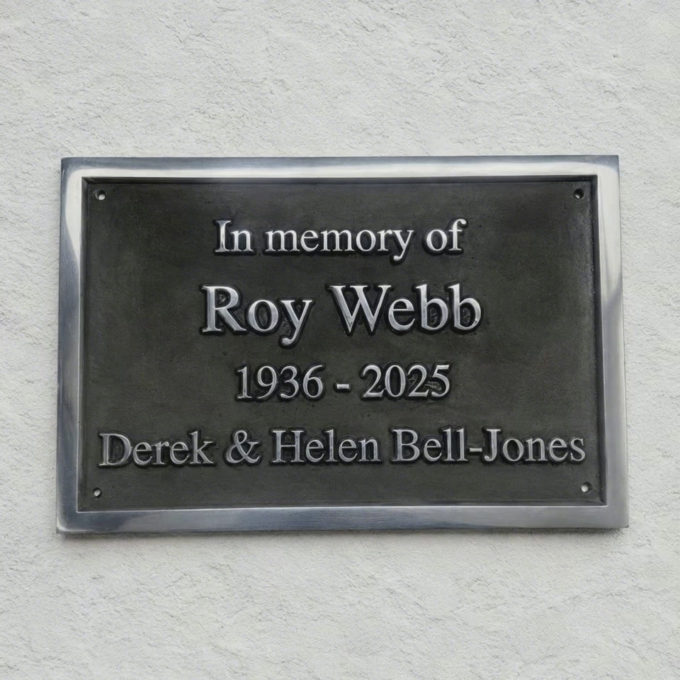 A rectangular dark metal memorial plaque mounted on a textured white wall, with raised silver text reading 'In memory of Roy Webb 1936 - 2025 Derek & Helen Bell-Jones'.